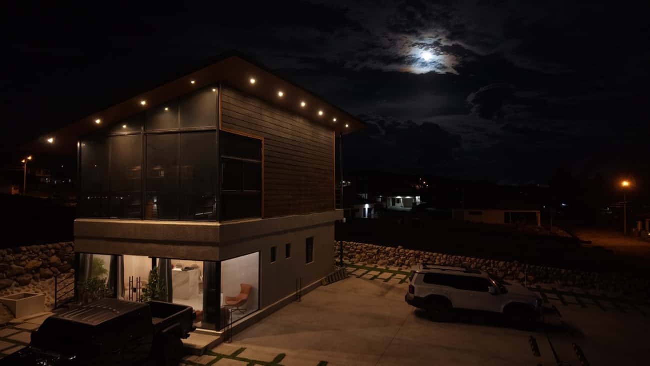 Cabaña iluminada de noche con parking privado al frente y luna sobre las montañas